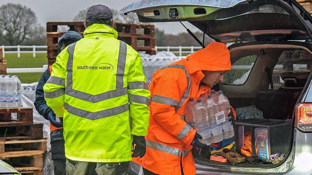 A man in a yellow hi-vis jacket with the word South East Water on the back. Another man, in an orange hi-vis jacket, is carrying a slab of water out of a car.