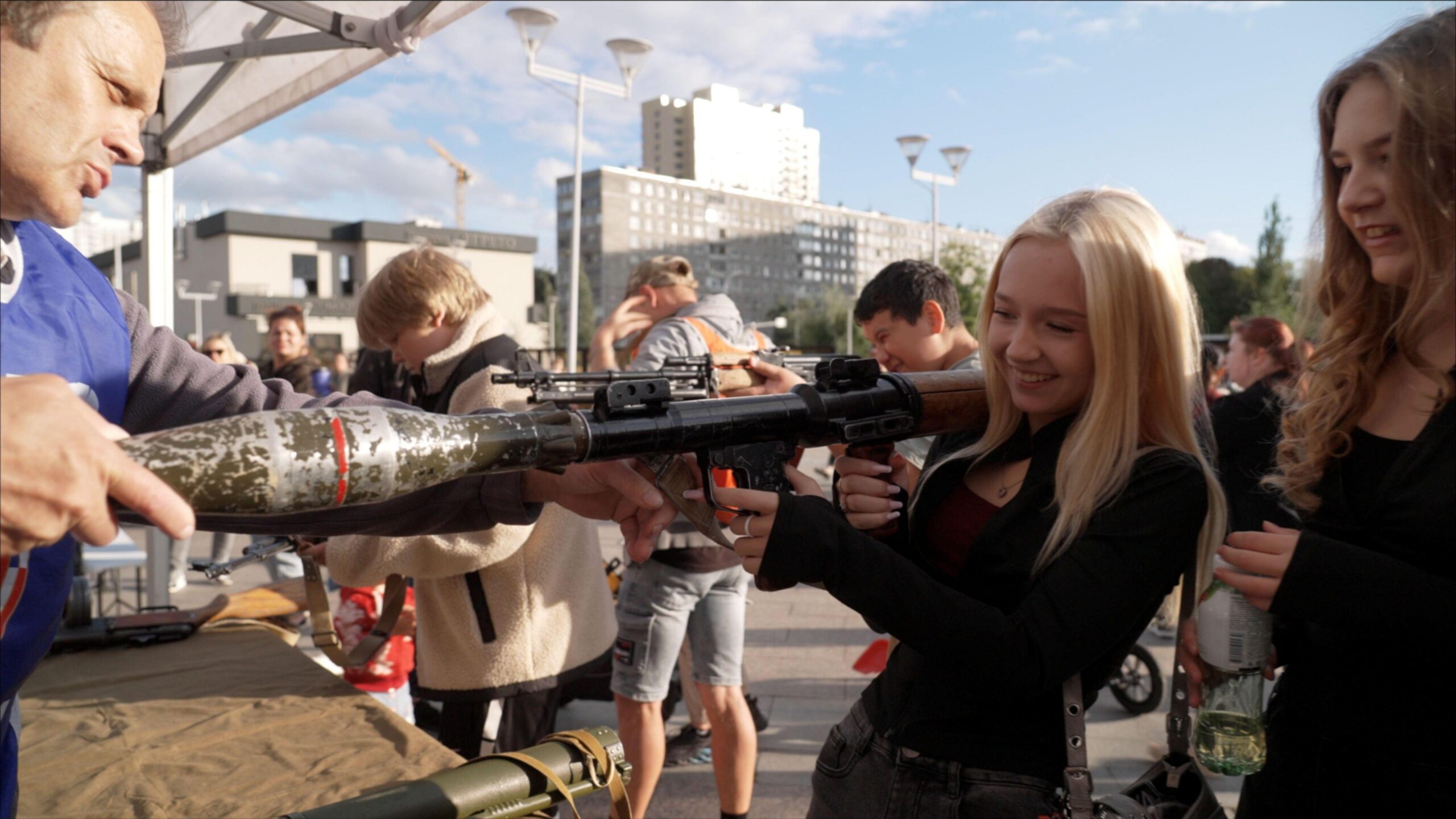 A blonde Russian child in a black top smiles as she holds a rocket launcher, at a weapons demonstration in Moscow