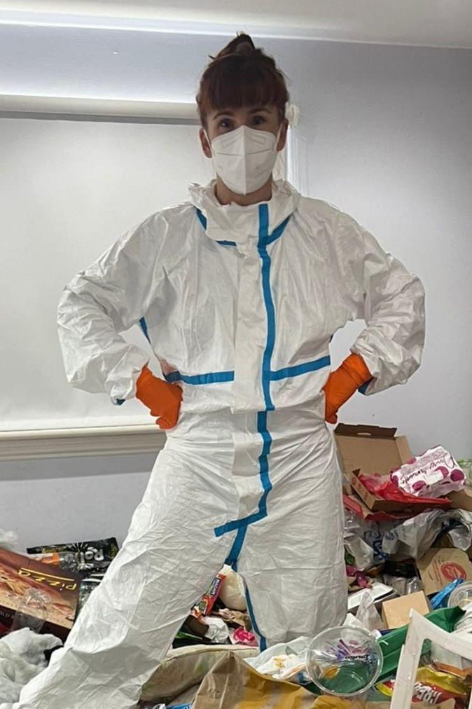 A woman standing on a pile of rubbish inside a house. She has ginger hair tied back and a fringe. She is wearing a white hazmat suit, a white face mask and orange gloves.