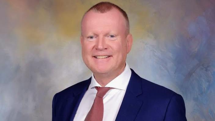 Professional studio portrait of Dennis Bacon wearing a navy suit, white shirt and muted red tie, facing the camera against a softly blended blue, grey and beige background. The image is sharply focused with even lighting and a formal corporate style.