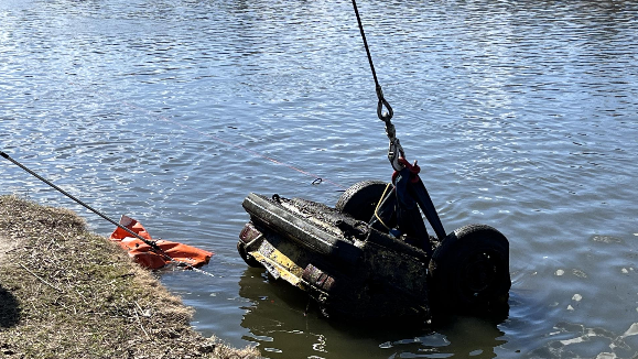 A heavy rope and chain drag an upside-down car from the shores of a river
