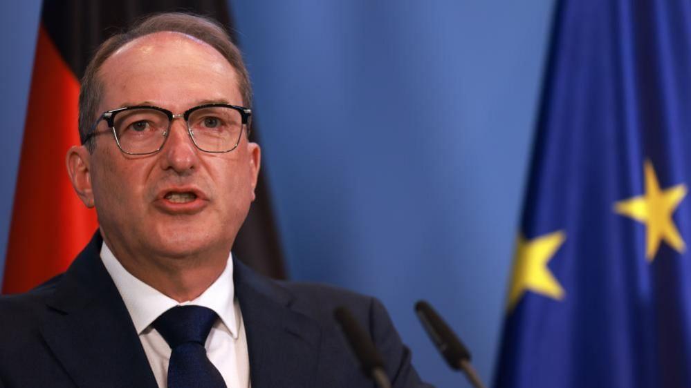 An older man with a receding hairline and glasses wears a dark suit and tie.