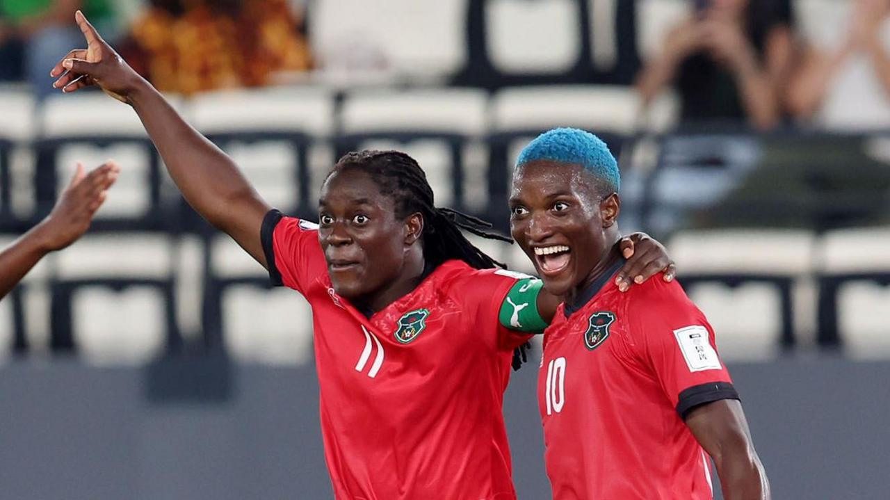 Tabitha Chawinga raises her right arm in celebration and puts her left arm around the shoulder of Temwa Chawinga, who has dyed blue hair and a wide-mouthed smile on her face. Both players wear red Malawi shirts with black trim - Tabitha with a green captain's armband and a white number 11 on the front and Temwa with a number 10 on the chest. The pair are visible from stomatch up and a sparsely-populated stand can be seen out of focus in the background