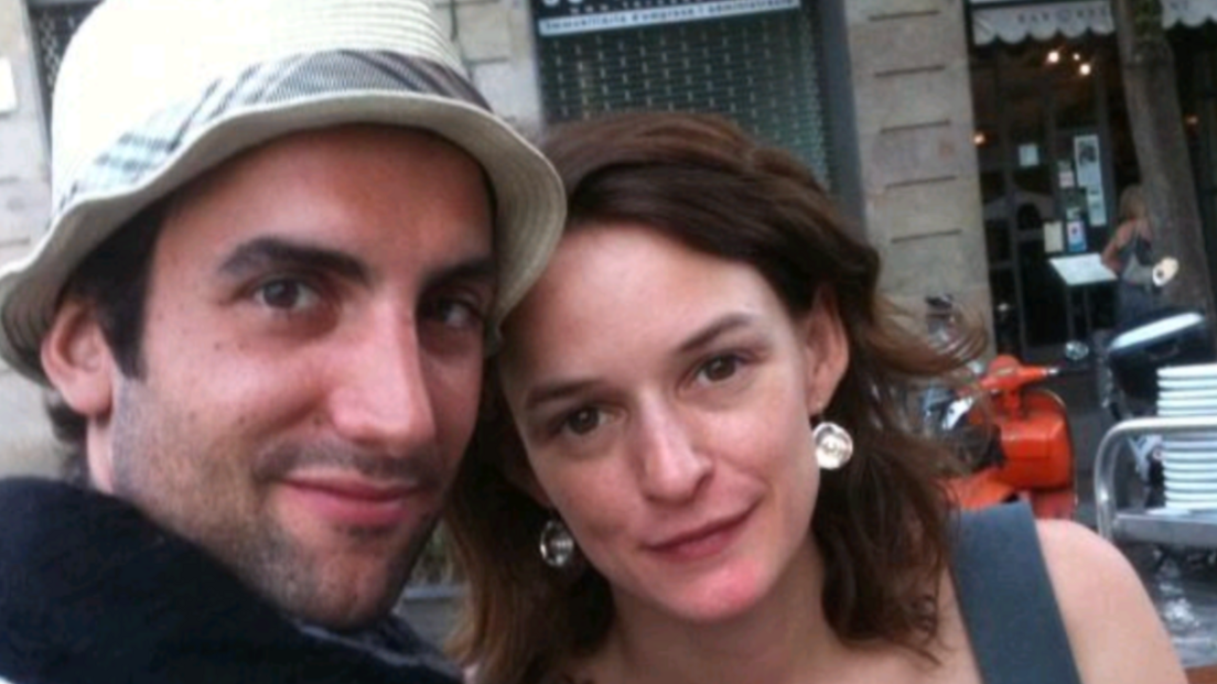 A close-up photograph shows a man and a woman - both with brown hair - posing together outdoors and looking toward the camera. The man wears a white hat. The backdrop is a city street with a building façade, outdoor seating and an orange scooter visible in the background.