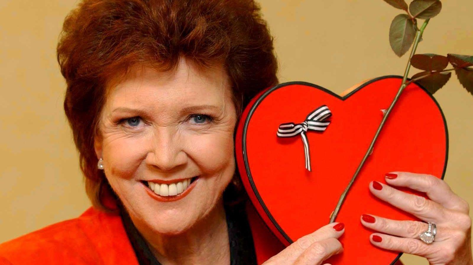 Cilla Black smiling and holding up a love heart while host of ITV show Blind Date