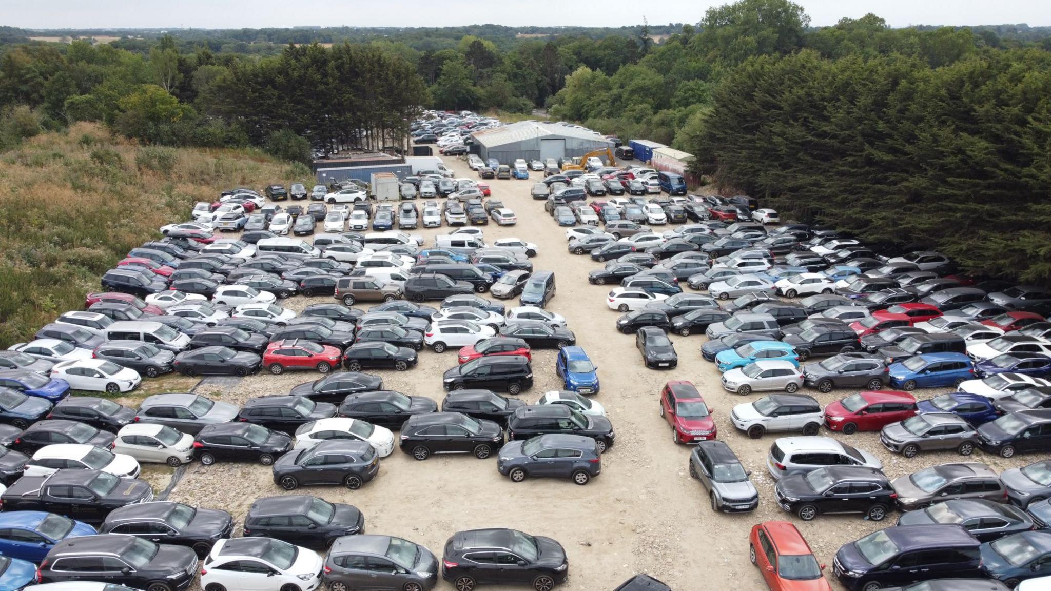 An aerial view of a large open-air car park. There are hundreds of cars lined up bumper to bumper, parked on gravel. There is a grey warehouse in the distance. The car park is surrounded by trees.