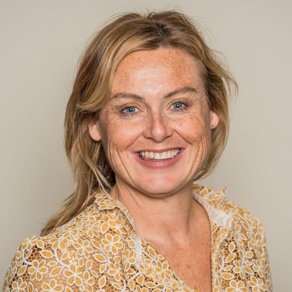 Person with shoulder-length hair facing the camera against a plain background, wearing a patterned lace blouse with a floral design and a collar. The image is a head-and-shoulders portrait, with the upper body and face clearly visible. No other objects or text are present in the image.