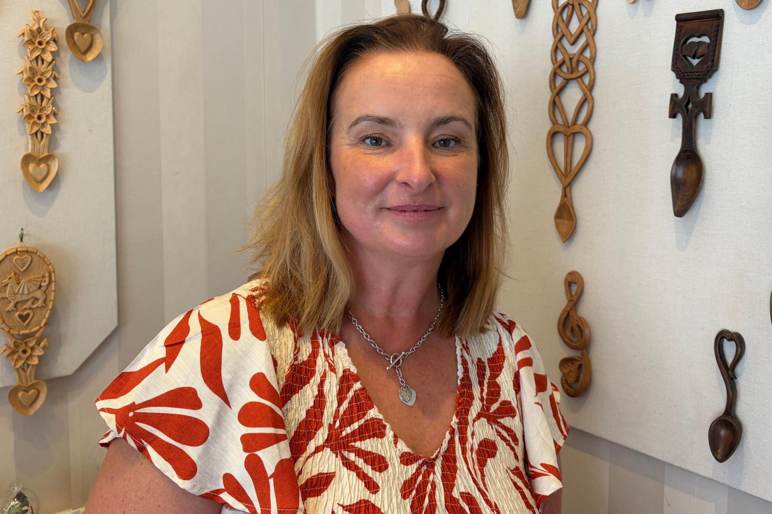 Jane, who has light brown hair and wears a floral white and red top, with love spoons attached to a wall in the background.
