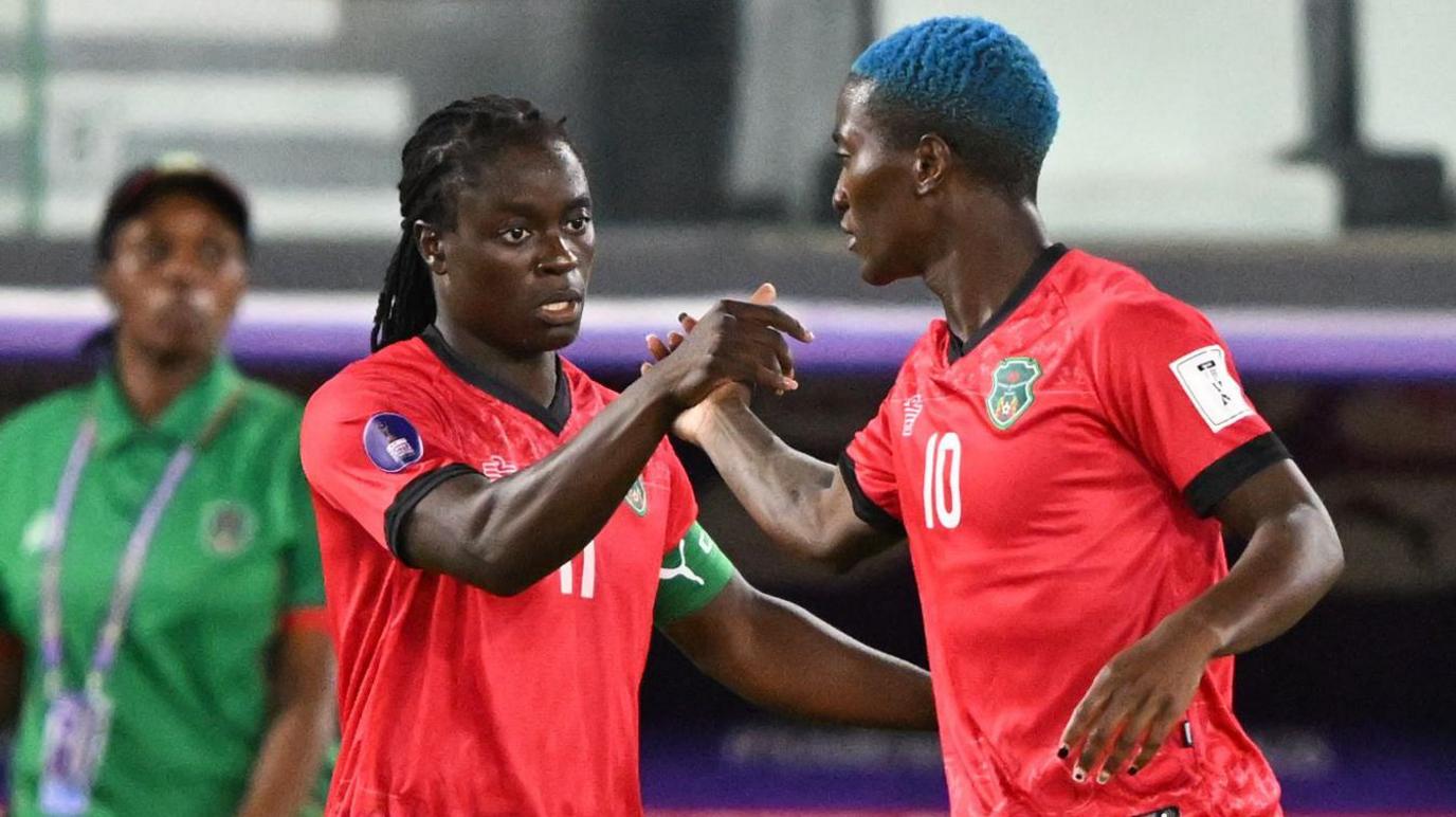 Tabitha Chawinga holds her right hand forward to clasp the hand of Temwa Chawinga, who is side-on and facing away from the camera. Both players wear red Malawi shirts with black trim - Tabitha with a green captain's armband and a white number 11 on the front and Temwa with dyed blue hair and a number 10 on the chest. A coach in a green shirt, a dugout and sparsely-populated stand can be seen out of focus in the background