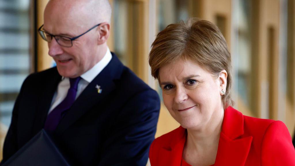 John Swinney and Nicola Sturgeon walking through the corridors of the Scottish Parliament. He wears black-rimmed spectacles, a blue suit, a purple tie and a white shirt. She is dressed in a red blazer and smiles at the camera.