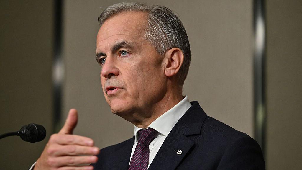 Prime Minister Mark Carney speaking at a podium. He has short grey-black hair and is wearing a black suit with a burgundy tie.