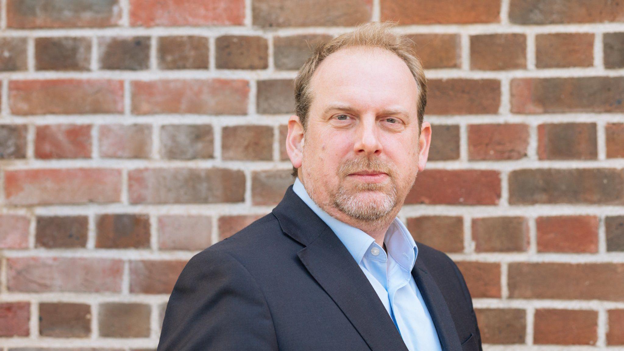 Carr-West, with greying red hair looks into the camera. He is pictured as a head and shoulders shot, wearing a blue suit and a sky blue shirt. In the background is a brick wall.