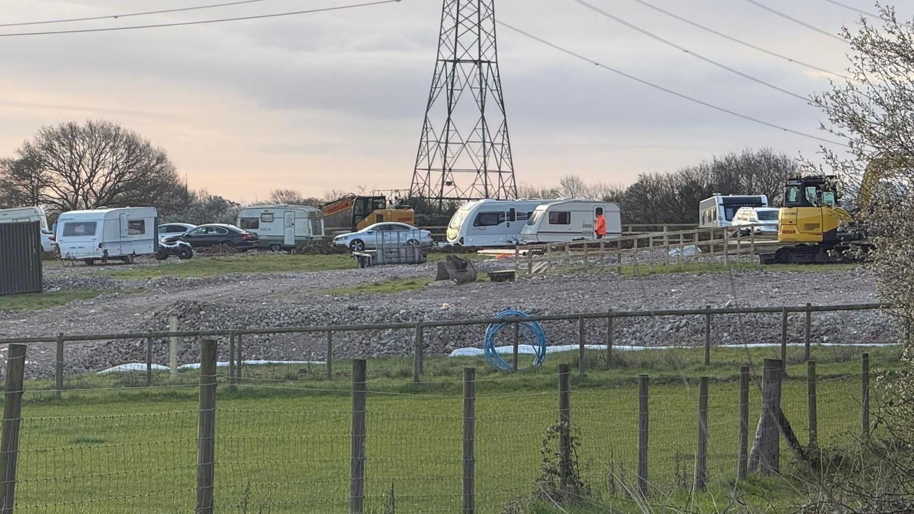 A view of the field. About six caravans are parked on hardstanding along with other vehicles and yellow diggers. There is a pylon in the middle of the field.