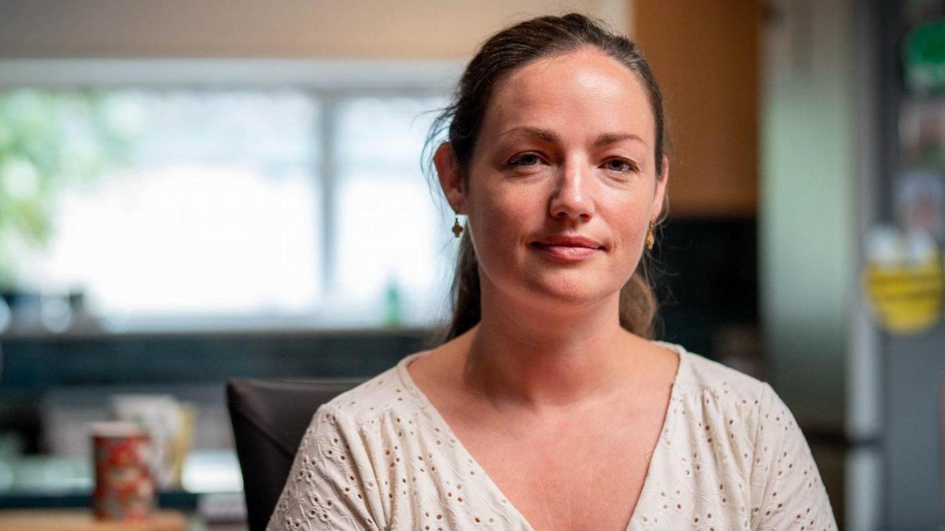 A head and shoulders image of Cat Razzell. She is looking directly at the camera. Her dark hair is tied back off her face. She is wearing a cream broderie top and dangly earrings. Behind her, slightly blurred, is a room - possibly a kitchen..