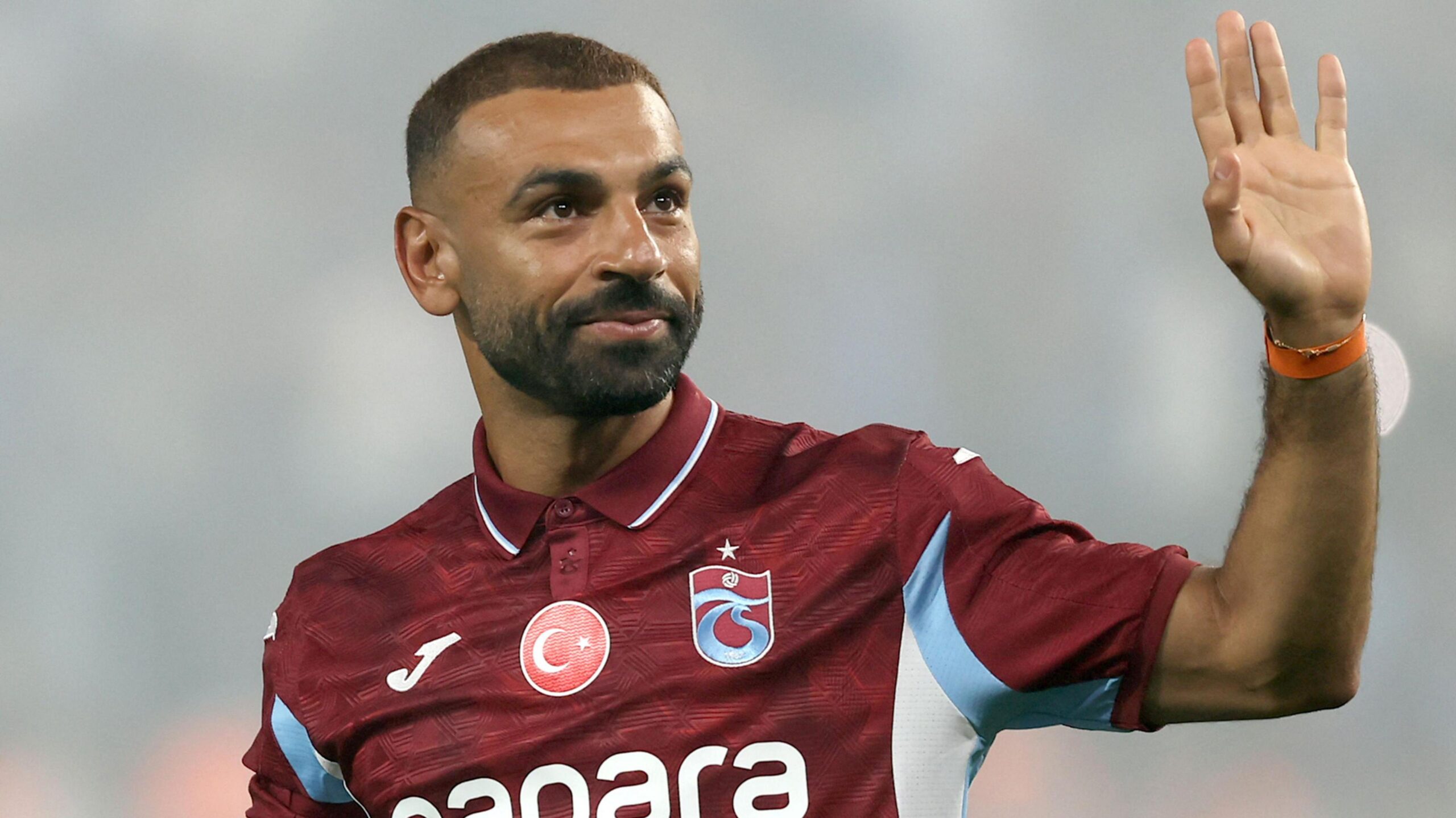 Mohamed Salah, visible from chest up wearing a claret Trabzonspor polo shirt with white and sky blue trim and a circular Turkish flag in the upper centre of the chest, raises his left hand to wave while showing a closed-mouth smile on his face