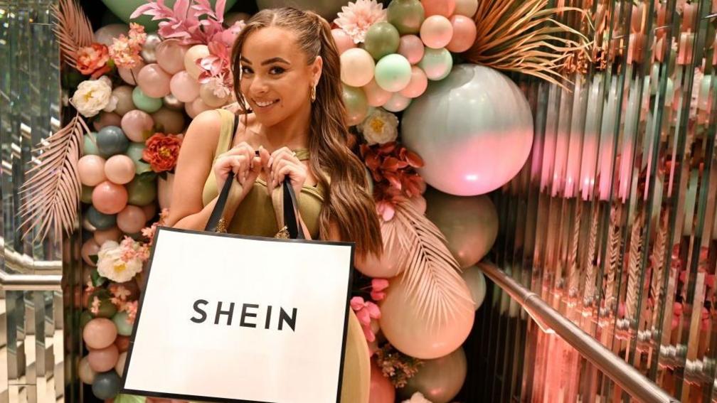 A woman poses for a picture while holding a Shein-branded shopping bag. She is surrounded by pastel-shaded balloons in a room.