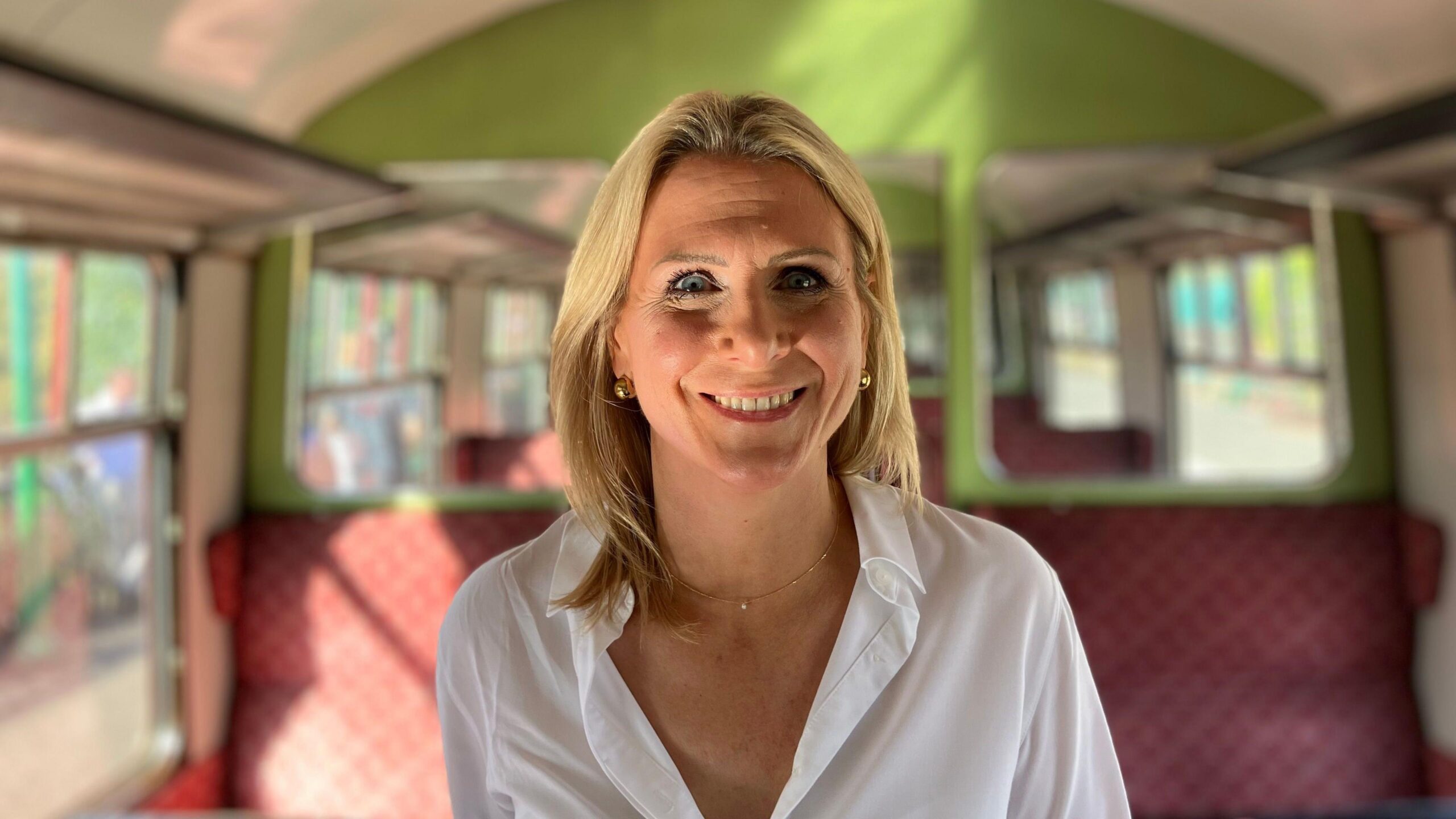 Erica Gilson has blonde shoulder-length hair and is wearing a white open-necked shirt. She is standing in the vintage train carriage, and is grinning.