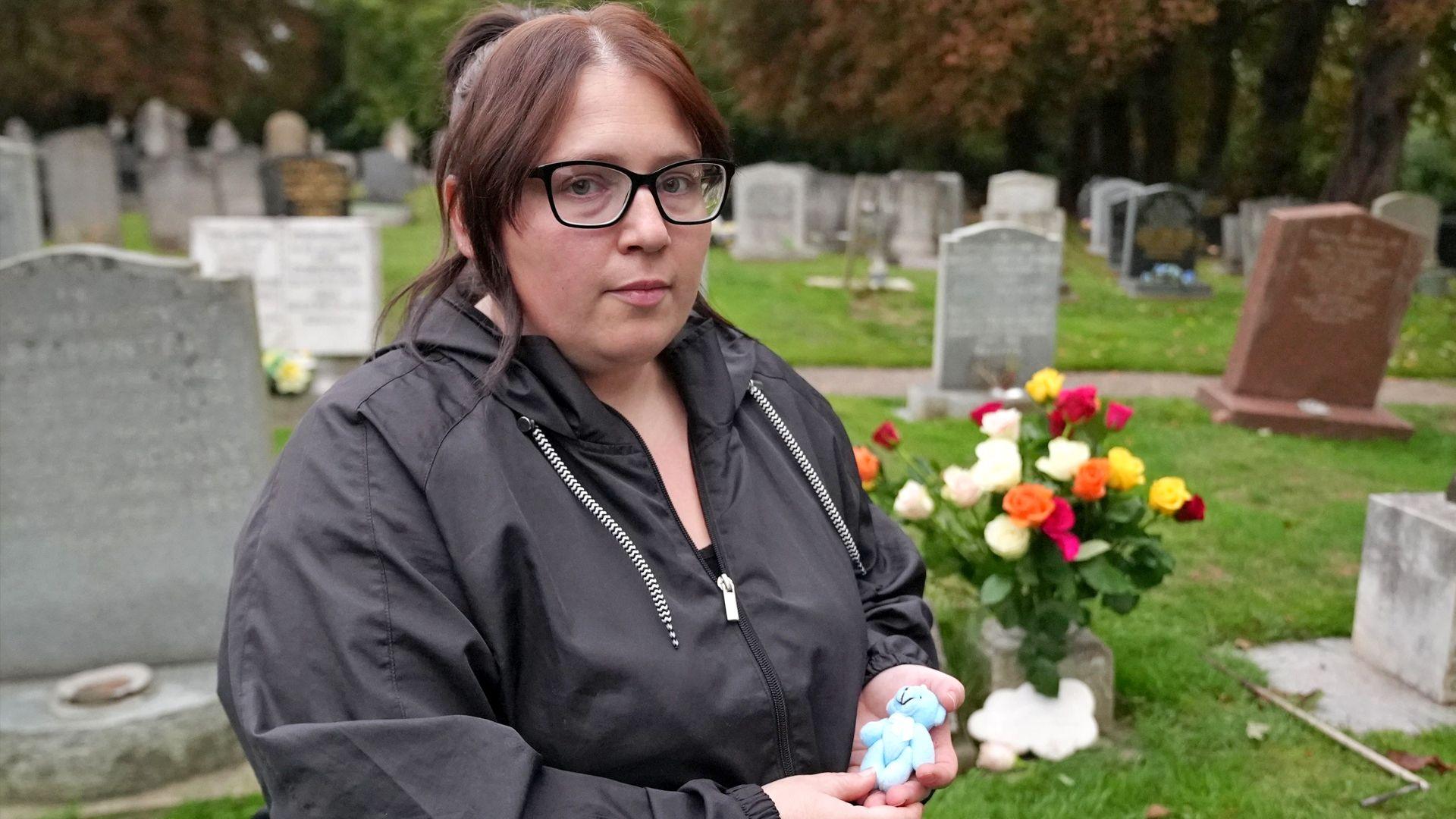 Jasmine Beverley wearing a black coat and black glasses holding her premature baby's small blue teddy bear. She is stood in front of her son's grave which is full of coloured roses.
