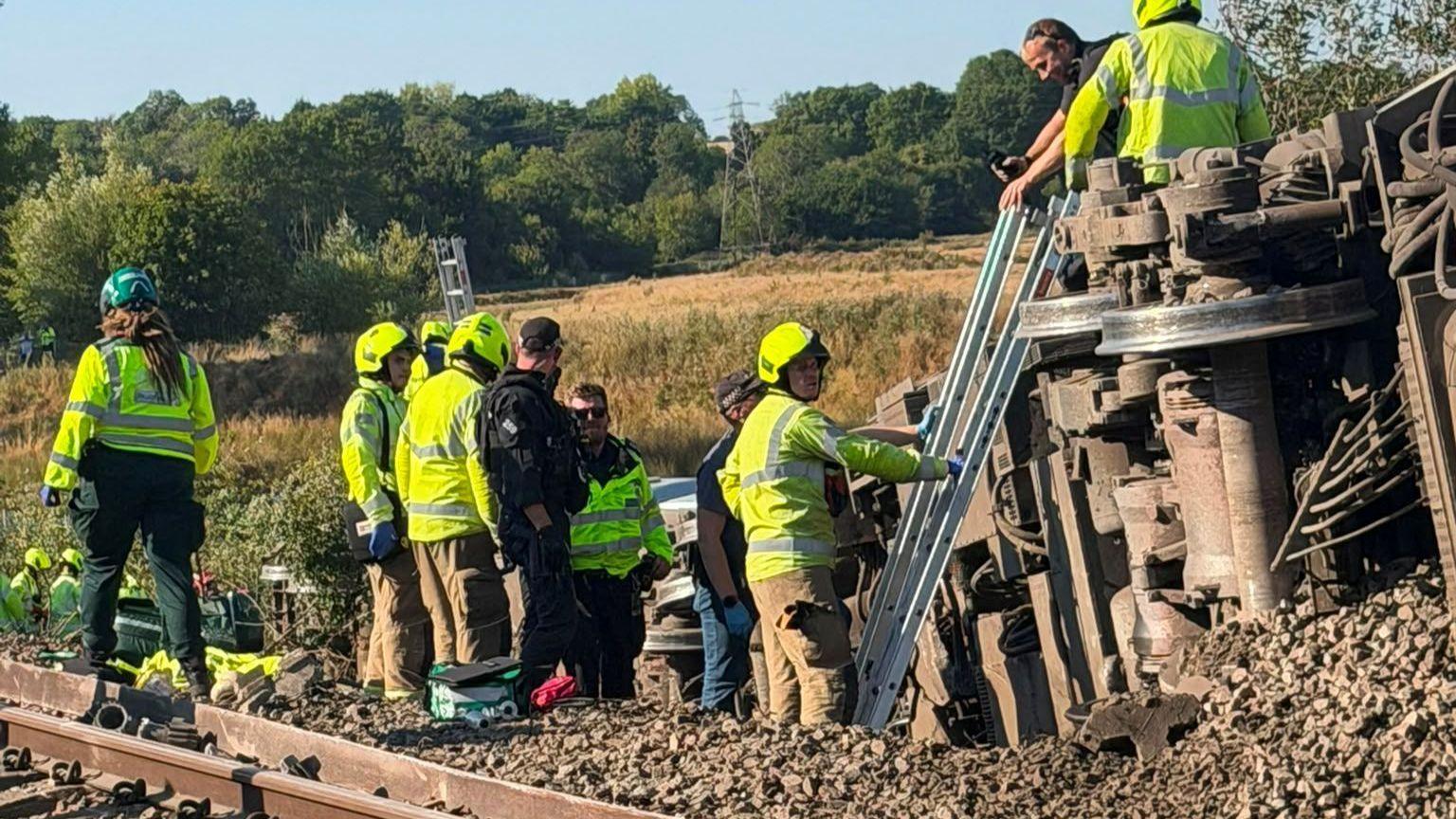 Emergency responders and rail workers stand beside a derailed train carriage resting on its side. Ladders have been placed against the carriage as crews work on the railway line surrounded by open countryside.