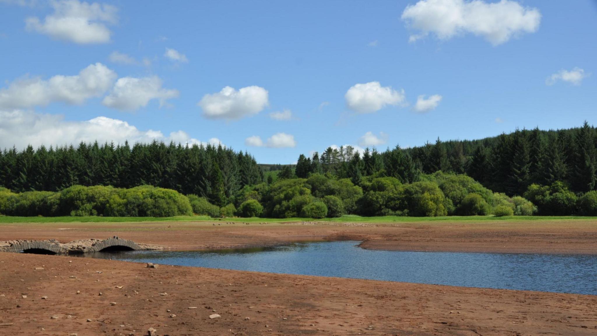 A dried out reservoir bed with a small amount of blue water and an old stone bridge.  Green grass, trees and blue sky with clouds are in the background.