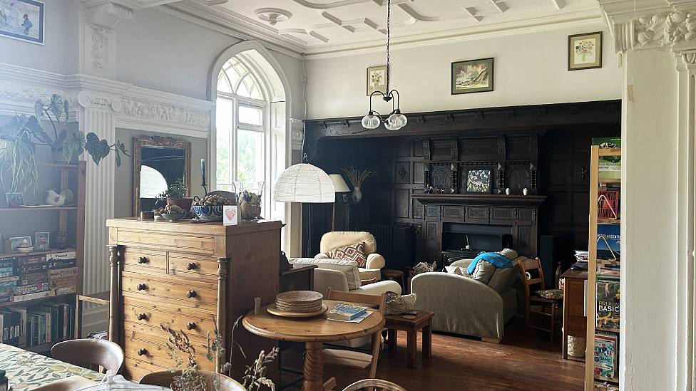 The room has very high ceilings with plaster features on it and carved white pillars at either side of a sitting area and a high arched window painted white with fanlight effects at the top. On the back wall is a dark wooden frieze with a mantelpiece and fireplace, giving a mock-Tudor appearance. There are a number of sofas and armchairs and a tall wooden chest of drawers in the middle of the room with a round coffee table next to it, making a demarcation between the living area and a dining area. The edge of a dining table and chairs can be seen in the bottom left corner of the photo. there are bookcases either side of the room with multiple books, games and plants on them. High on the walls above the line of the dark panelled area and a white wooden horizontal panel are a number of framed paintings. There are two suspended lampshades and a standard lamp in the corner by the window.