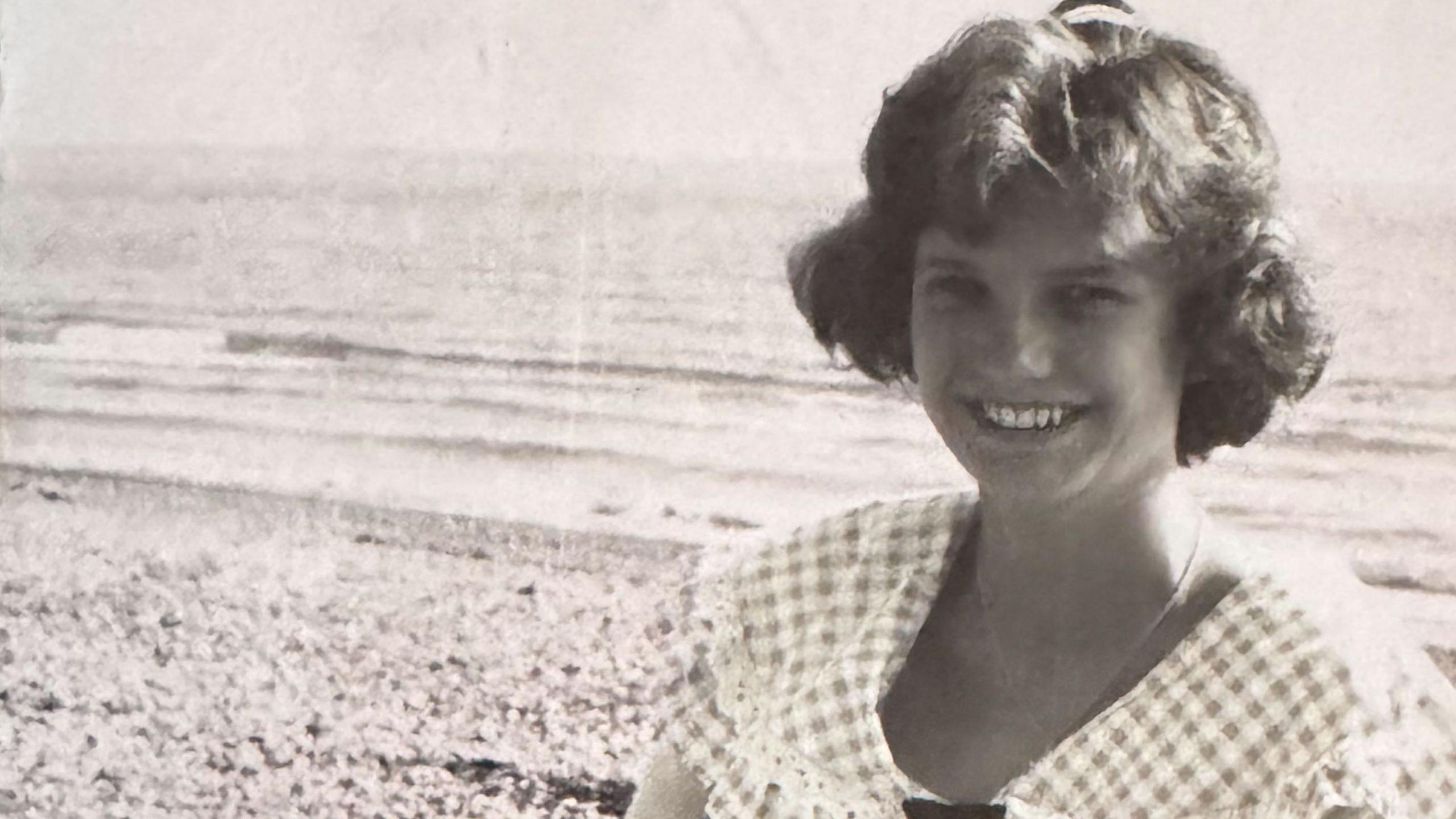 A black and white picture of a young woman in a gingham dress. She is sitting on a pebble beach with the sea behind her.