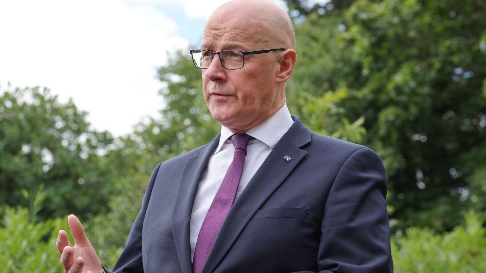 John Swinney, who is bald with glasses, speaks outdoors with leafy green trees in the background. He is wearing a business suit.