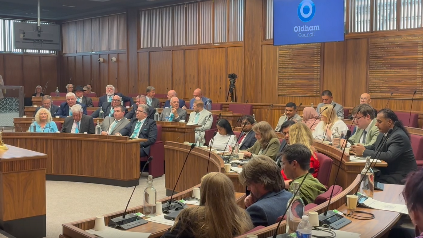 Oldham Council's chamber: a wood-panelled room with red seats and wooden desks in a u-shape around a central podium. There are people sitting in most of the seats. At the back is a screen with the Oldham Council logo.