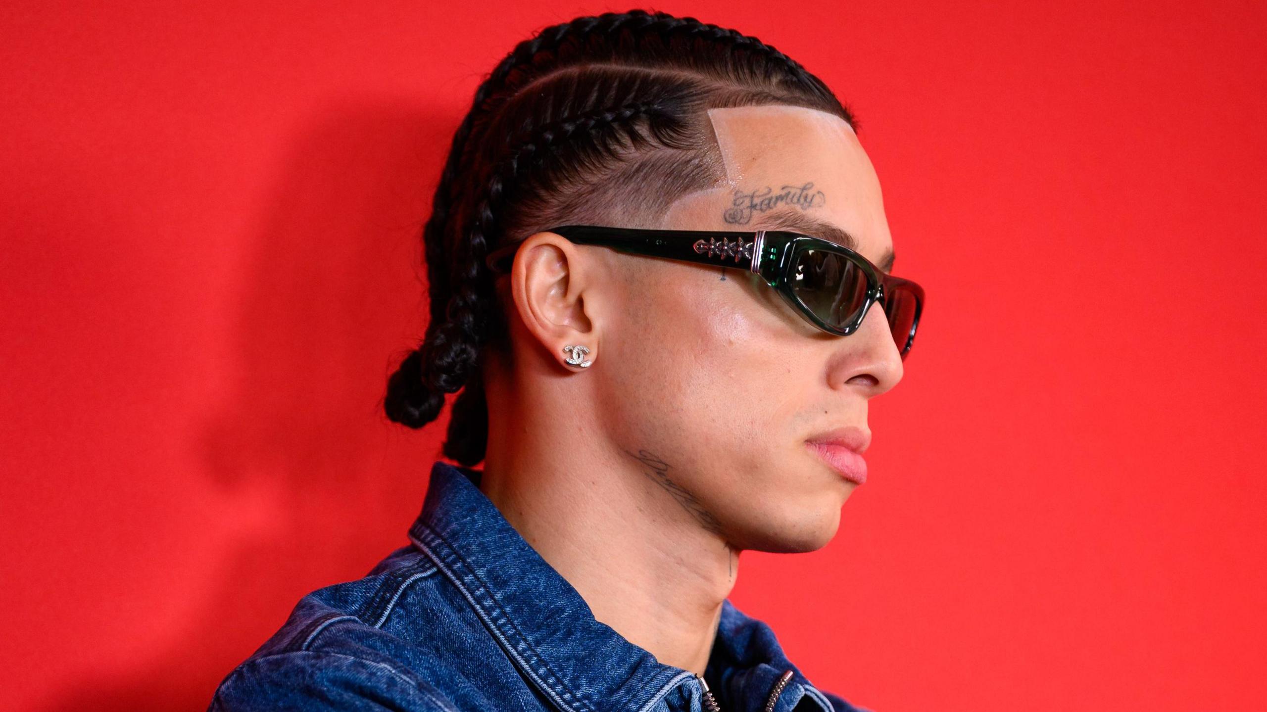 Central Cee pictured at the GQ awards in 2024. He wears a denim jacket and sunglasses, the tattoo 'family' above his right eye visible in the side-profile shot. He's pictured against a red backdrop.