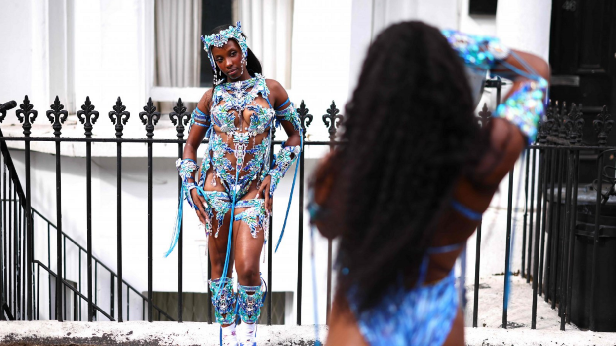 A woman in a heavily-beaded blue costume stands in front of a metal railing and poses for a photograph as another woman in a blue costume takes a picture.