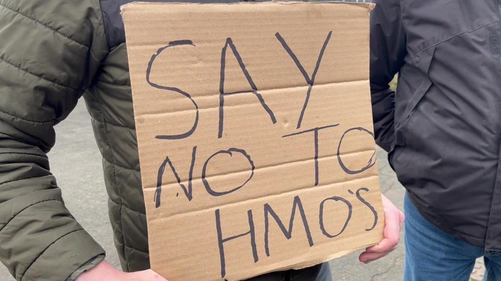 A campaigner holds a 'Say No To HMOs' sign written on cardboard. The person is wearing a green insulated jacket.