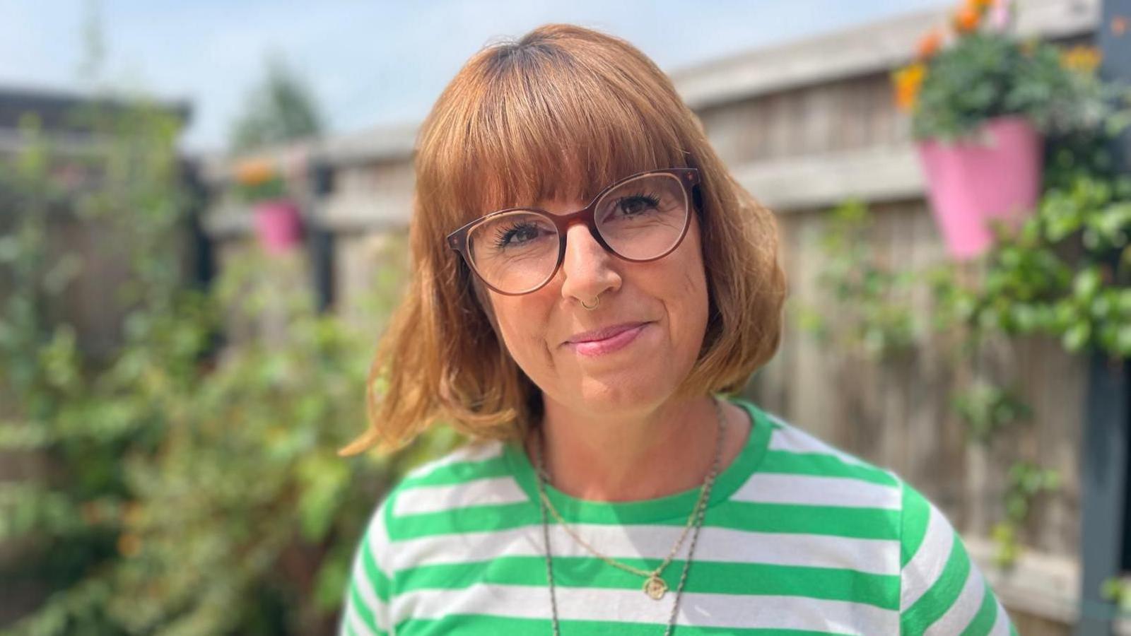 Catherine Warrilow with a light brown bob and fringe, wearing glasses and a green and white horizontal striped t shirt in a garden