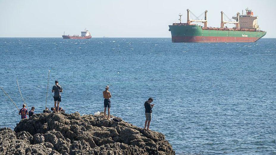 A group of men are seen from the back on a rocky outcrop with the blue sea and large ships in the background, on 22 August, 2026 in Ceuta, Spain.