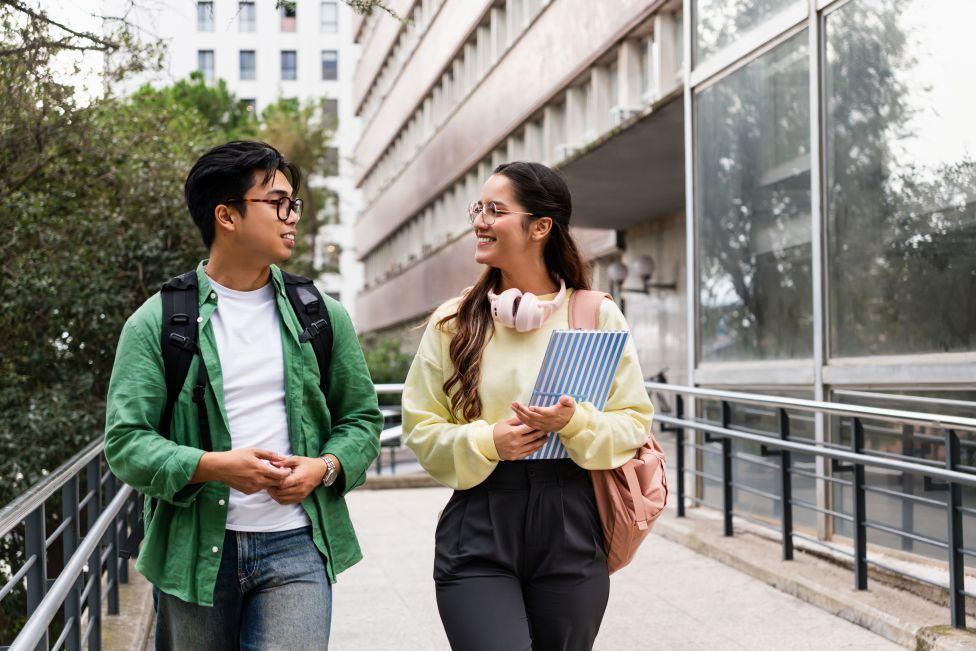 A man and a woman, both in glasses, smile and talk as the walk past a large university building