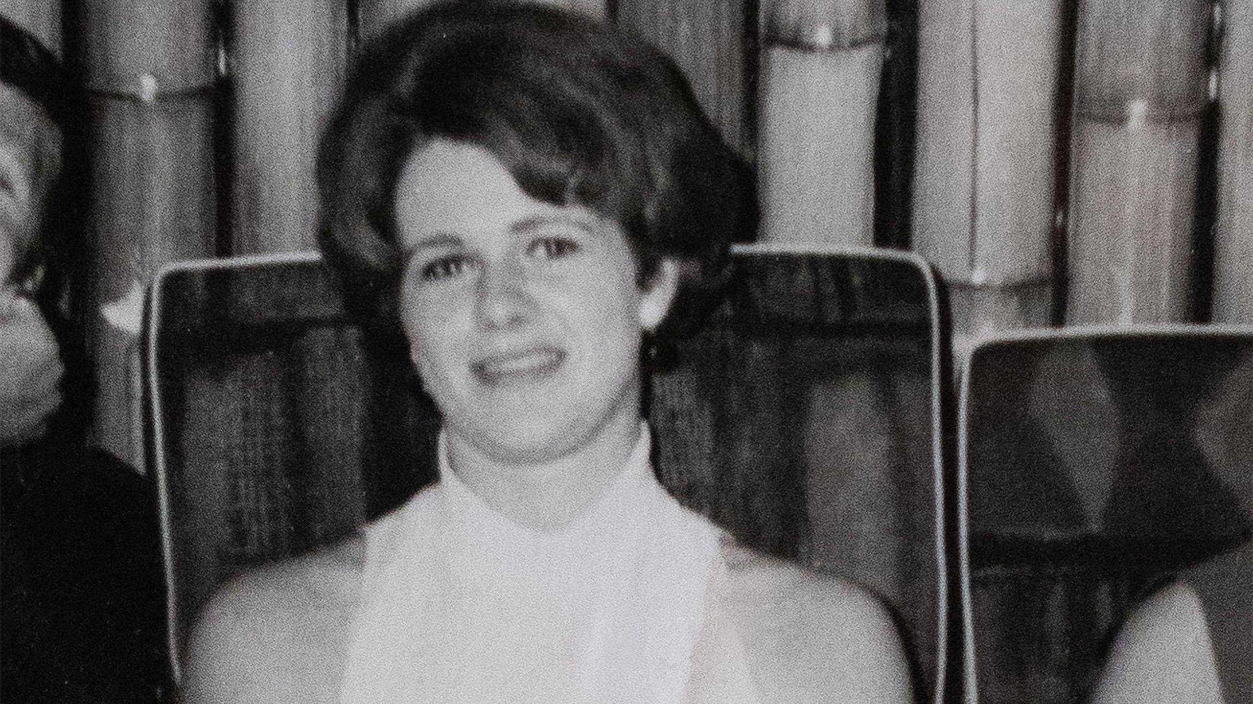A black and white picture of a young woman in a white, sleeveless top. She is sitting on a chair in front of curtain.