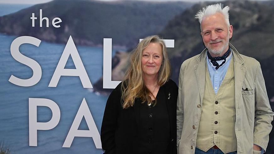 Win and Moth stood in front of a large screen which has a coastal scene and the words The Salt Path in big white letters. On the left is Winn with long blonde hair, wearing all black, and beside her is Moth with spiky white hair wearing a beige jacket and waistcoat, blue shirt and navy cravat. They are both looking at the camera and smiling.