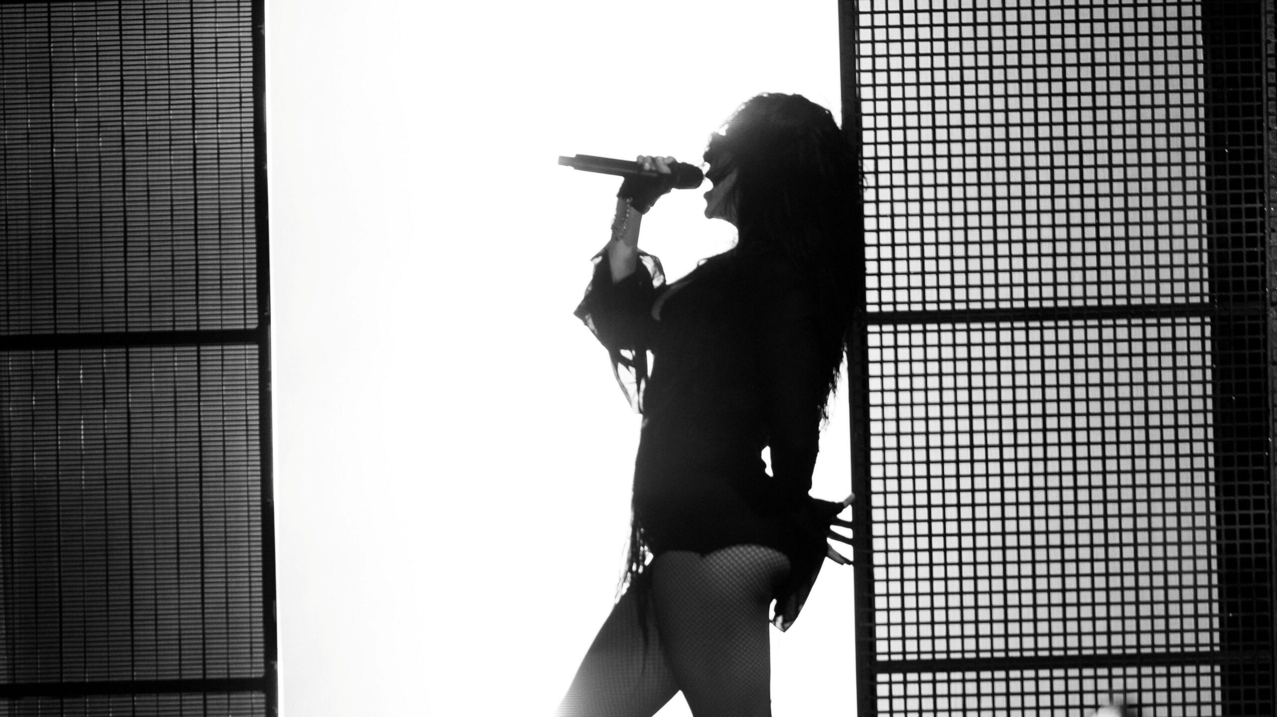 A black and white photo of Charli XCX leaning against a wall as she sings into a microphone. There is a bright white wall behind her which silhouettes her body.