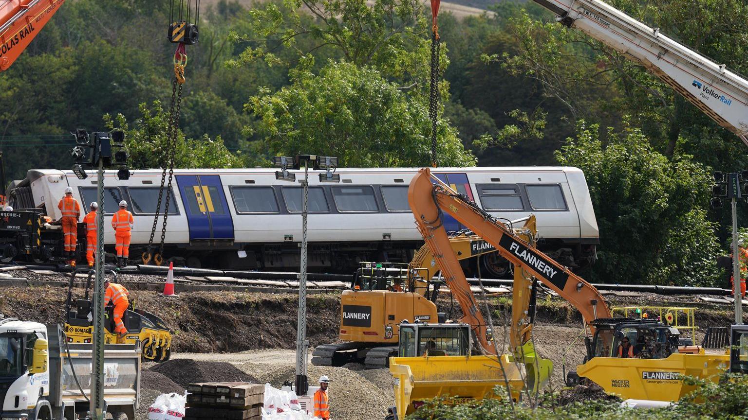Heavy machinery including cranes, excavators and construction vehicles is being used at the train derailment site near Lewes. A Southern train carriage is positioned beside the track while recovery crews work around the railway embankment and construction materials.