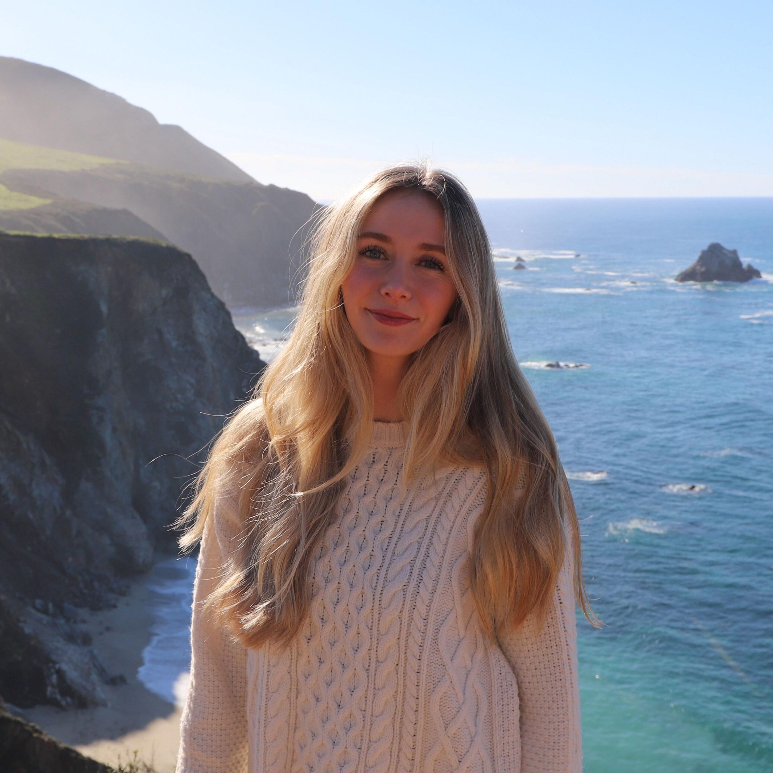 Person with long hair wearing a light-coloured knitted jumper standing in the foreground on a coastal cliff. Steep rocky cliffs and a sandy cove extend along the coastline behind them. The sea stretches into the distance with several offshore rock formations visible on the water. Clear skies and strong sunlight illuminate the coastal landscape.