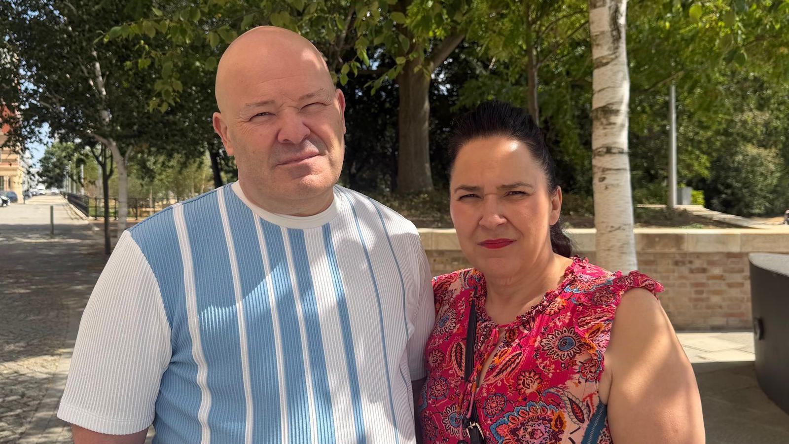 A man and a woman stand together on a pavement in front of a public garden. They have serious expressions on their faces. The man has a shaved head and wears a T-shirt with blue and white vertical stripes. The woman on the right has black hair, tied back and wears a red, pattered top.