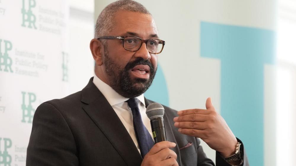 Sir James Cleverly, wearing a dark suit and tie, speaks into a handheld microphone at an event.