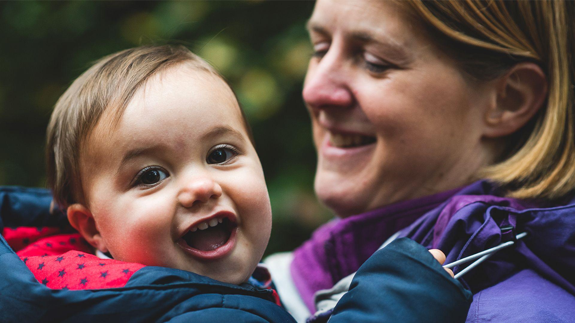 James, pictured as a young child, is held by his mother Laura in an outdoor setting, with greenery in the background.