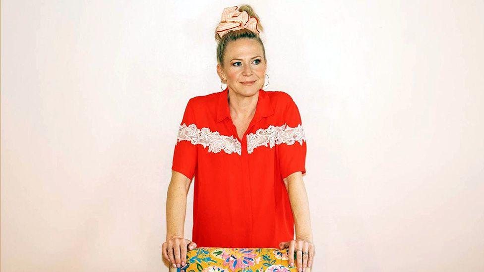 Kellie Bright is wearing a bright red shirt with white lace details, leaning forward with hands on a colourful floral-patterned surface, against a plain light background. Her hair is tied up with a pink accessory.