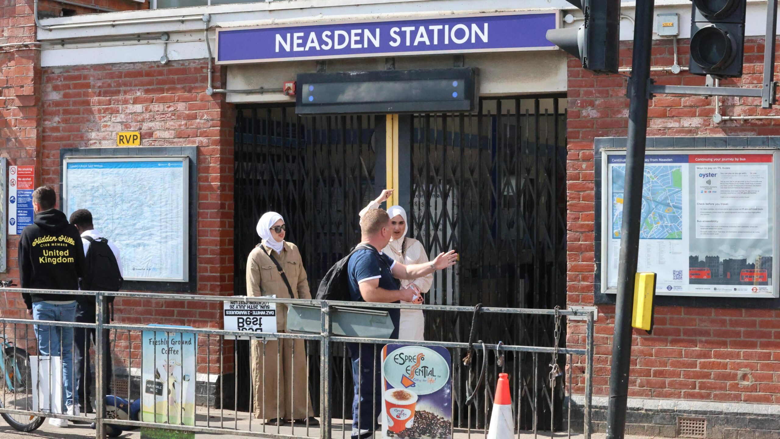 People gather outside the closed metal shutters of Neasden Station entrance, with one man gesturing towards the station signs while commuters wait on the pavement.
