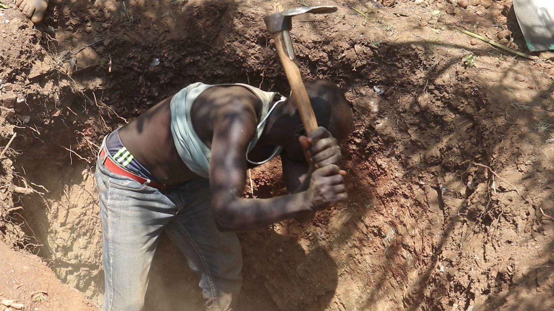 A man crouches in a trench with a hoe as he is about to bring it down to break up the earth