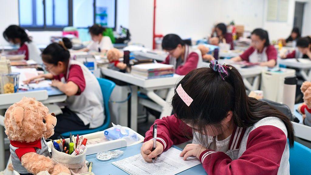 A teenager sits at a desk with her head down looking at a paper. She is writing on it with black pen. Stationery and a teddy bear sits on her desk. In the background are other teenagers at desks, all spaced out, in a classroom.