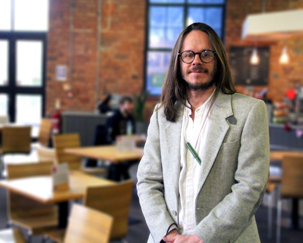 Darren Quelch, a man with long brown hair in a grey suit and glasses, stands in cafe and looks at the camera