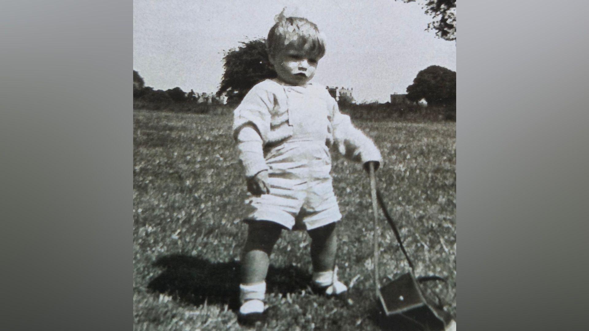 A black and white image of a toddler. The boy is playing outside. He is wearing dungarees with a long-sleeved top underneath, and socks and dark shoes.