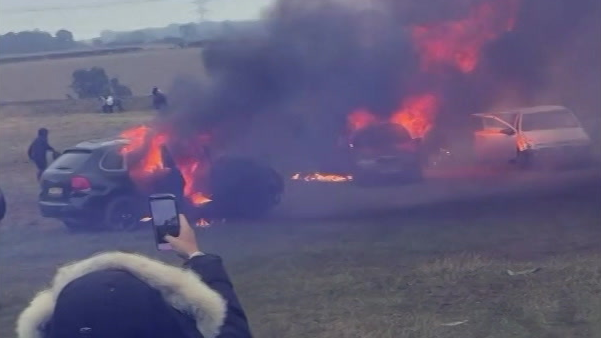 Cars ablaze with smoke billowing from their windows. There are people behind them running around and in the foreground is a person with their hood up, seen from behind, holding their phone up to capture the scenes.