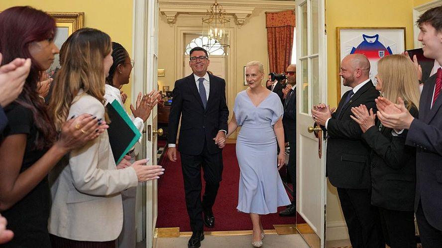 Staff applaud as Britain's new Prime Minister Andy Burnham and his wife Marie-France van Heel walk hand in hand into 10 Downing Street